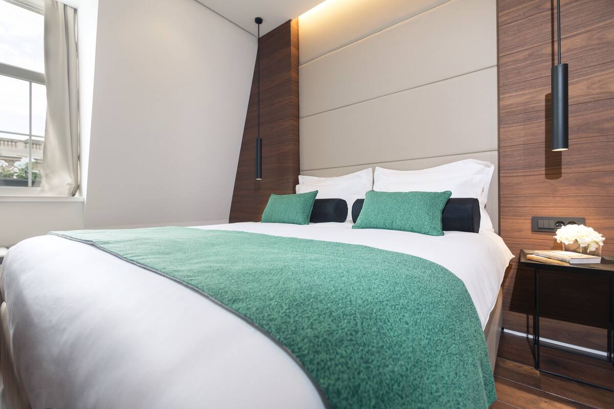 A modern bedroom with a large bed, green throw pillows, a wooden accent wall, and a bedside table with a book and flowers.