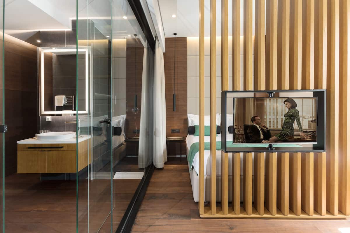A modern hotel room with a glass shower enclosure, a wooden vanity, a bed with a green and white striped bedspread, and a large mirror reflecting the room.