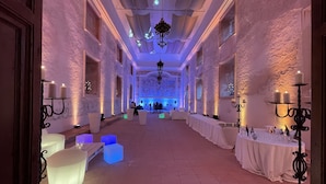 A spacious hall with white tables and chairs, a chandelier, and a stage at the far end.