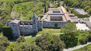 An aerial view of a historic building complex with a central courtyard, surrounded by lush greenery and a small parking area.