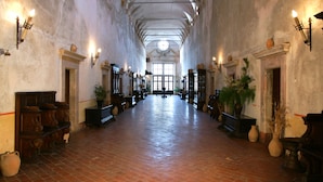 A long corridor with high ceilings and arched windows, flanked by wooden benches and wall-mounted sconces.