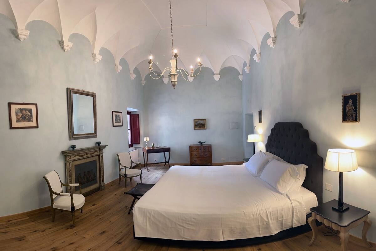 A bedroom with a large bed, a chandelier, a fireplace, and a desk with a chair.
