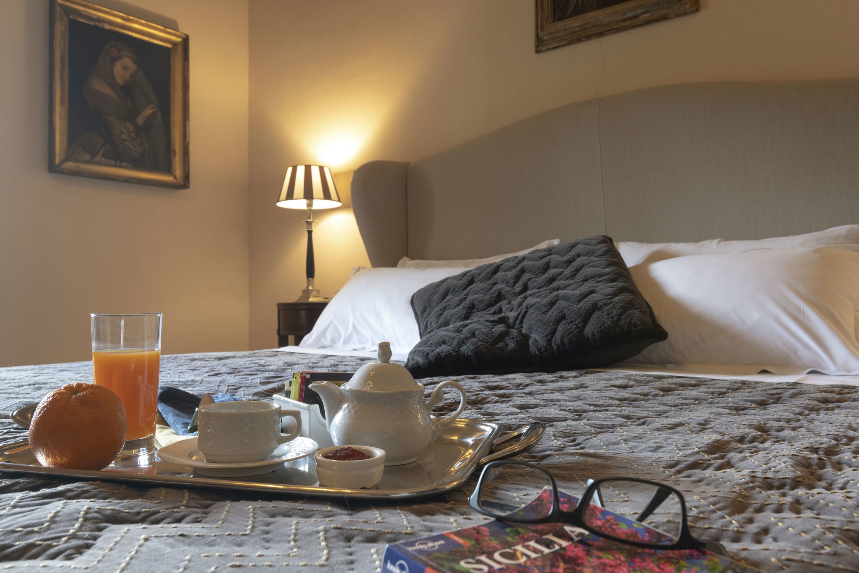 A neatly made bed with a pillow, a bedside lamp, and a tray with breakfast items including a muffin, a glass of orange juice, a teapot, and a cup.