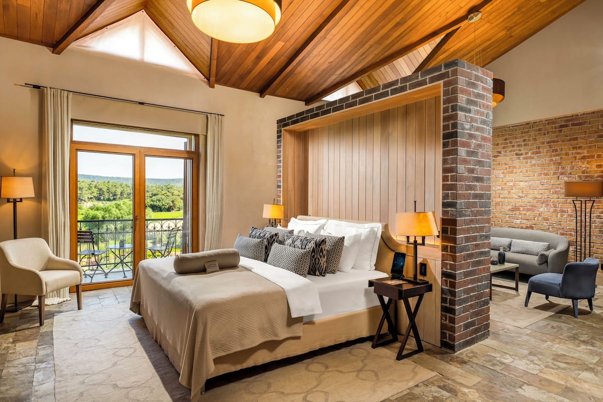 A spacious bedroom with a large bed, two bedside tables, a seating area with a sofa and armchairs, and a balcony with a view of the greenery.
