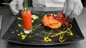 Un chef preparando un plato con salmón y tocino sobre un plato negro con salsa amarilla.