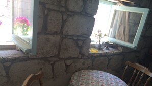 Quarto com paredes de pedra, uma pequena mesa coberta por uma toalha de mesa florida, uma cadeira e uma janela com cortina.