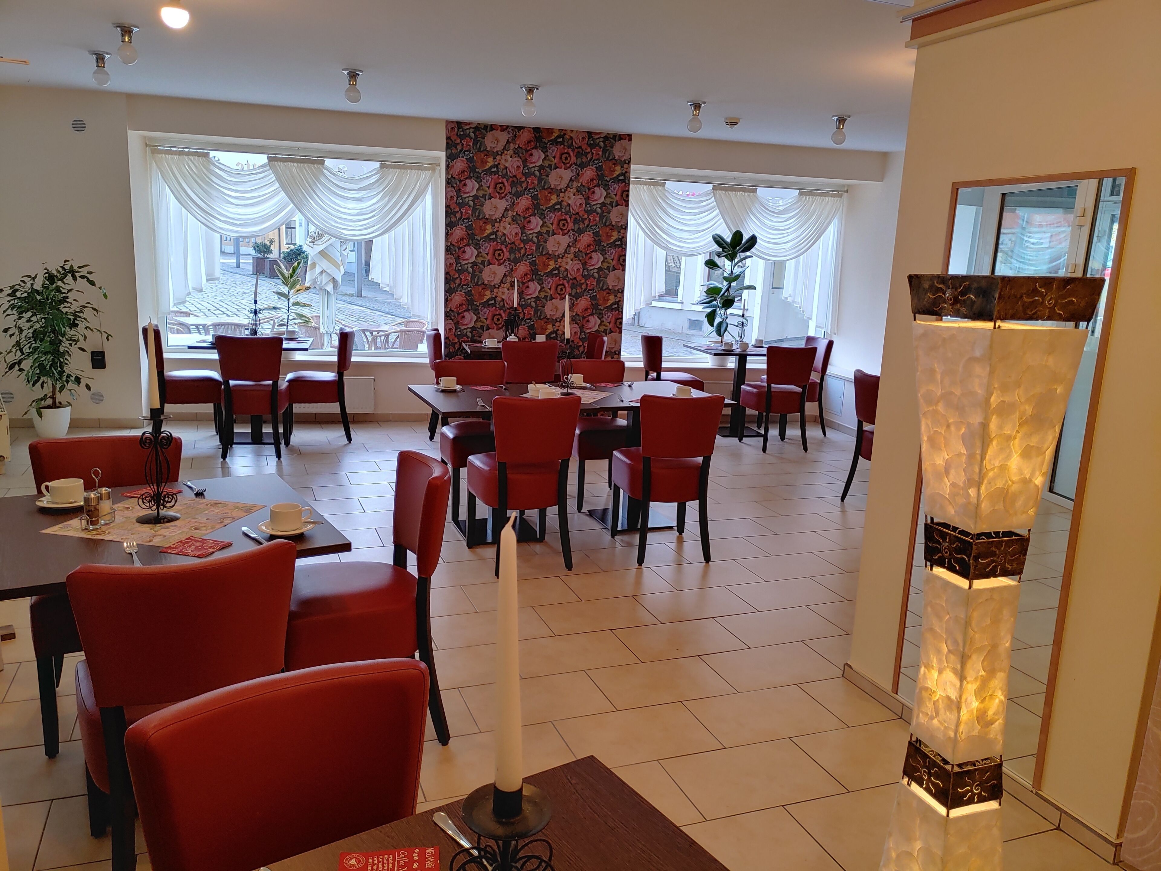 Un comedor amplio con sillas y mesas rojas, ventanas grandes con cortinas blancas y un estampado floral en la pared.