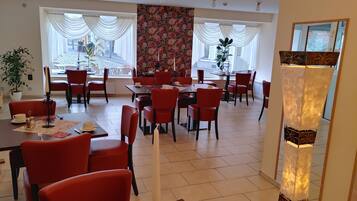 Un comedor amplio con sillas y mesas rojas, ventanas grandes con cortinas blancas y un estampado floral en la pared.