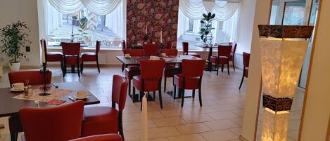 A spacious dining area with red chairs and tables, large windows with white curtains, and a floral wallpaper accent.