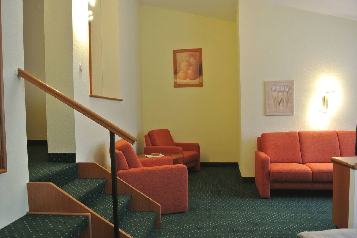 A hotel lobby with a staircase, orange armchairs, a sofa, and a painting on the wall.