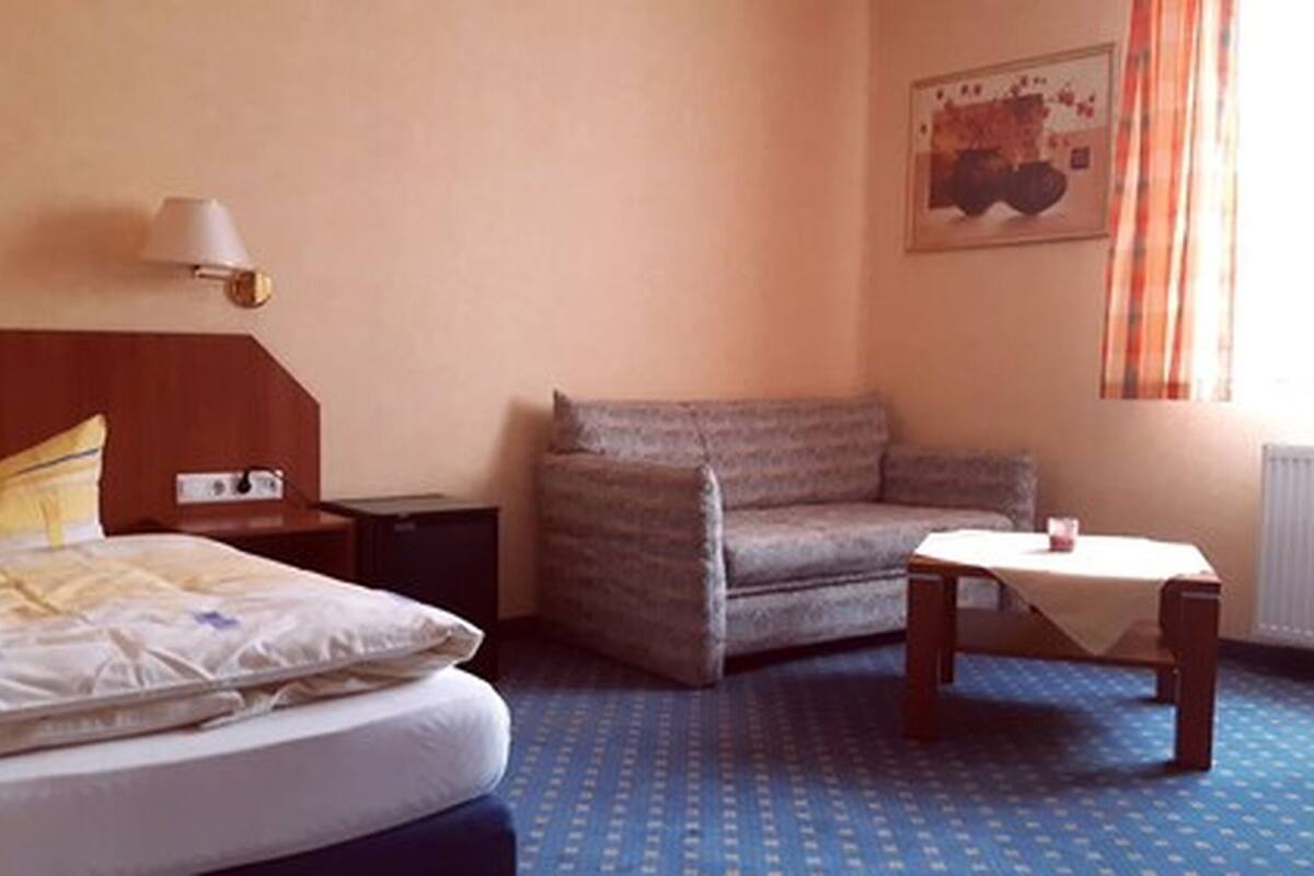 A hotel room with a bed, a chair, a small table, a lamp, and a painting on the wall.