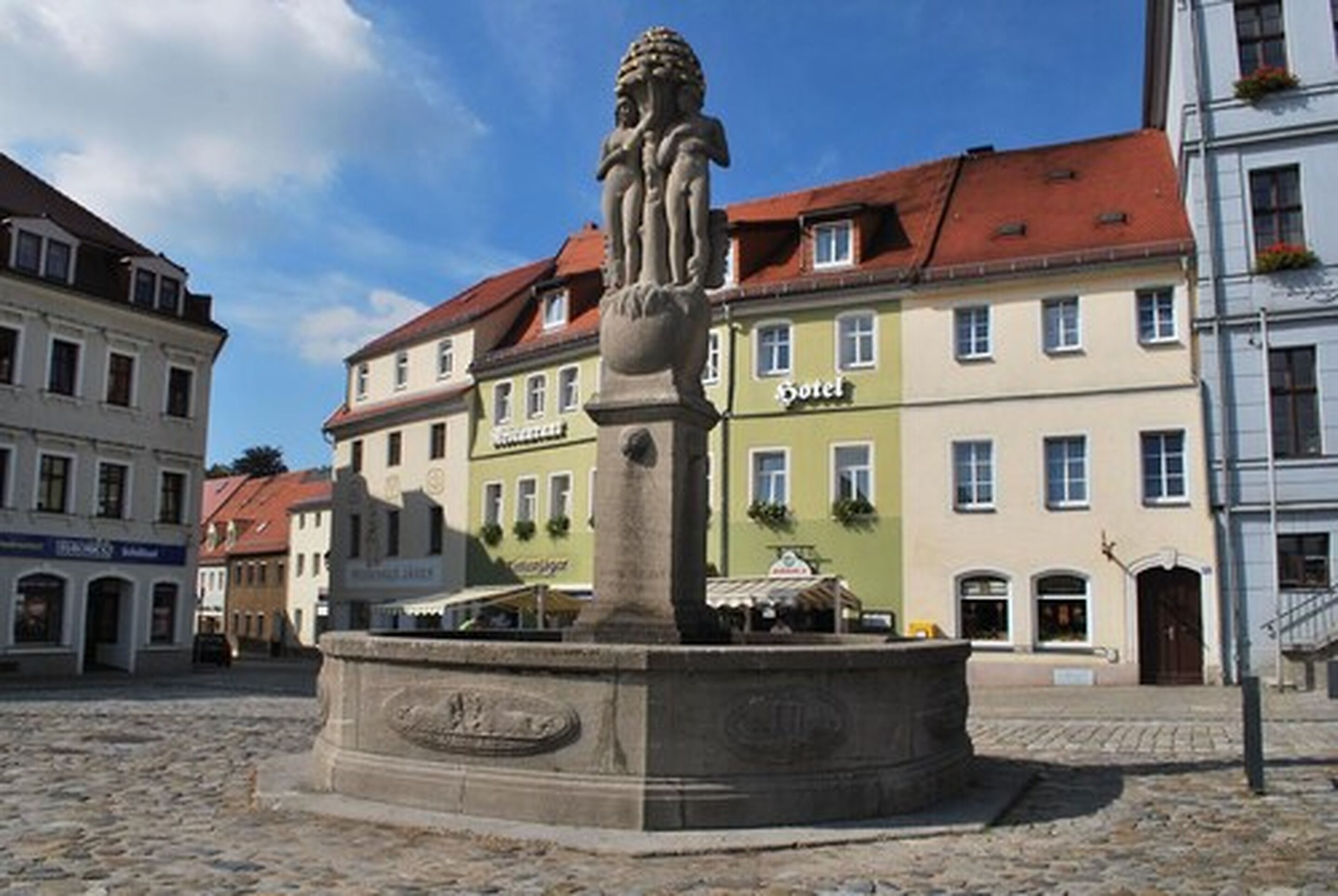 A stone fountain with a statue in the center, surrounded by cobblestone pavement. In the background, there are buildings with signs, including one that says "Hotel."
