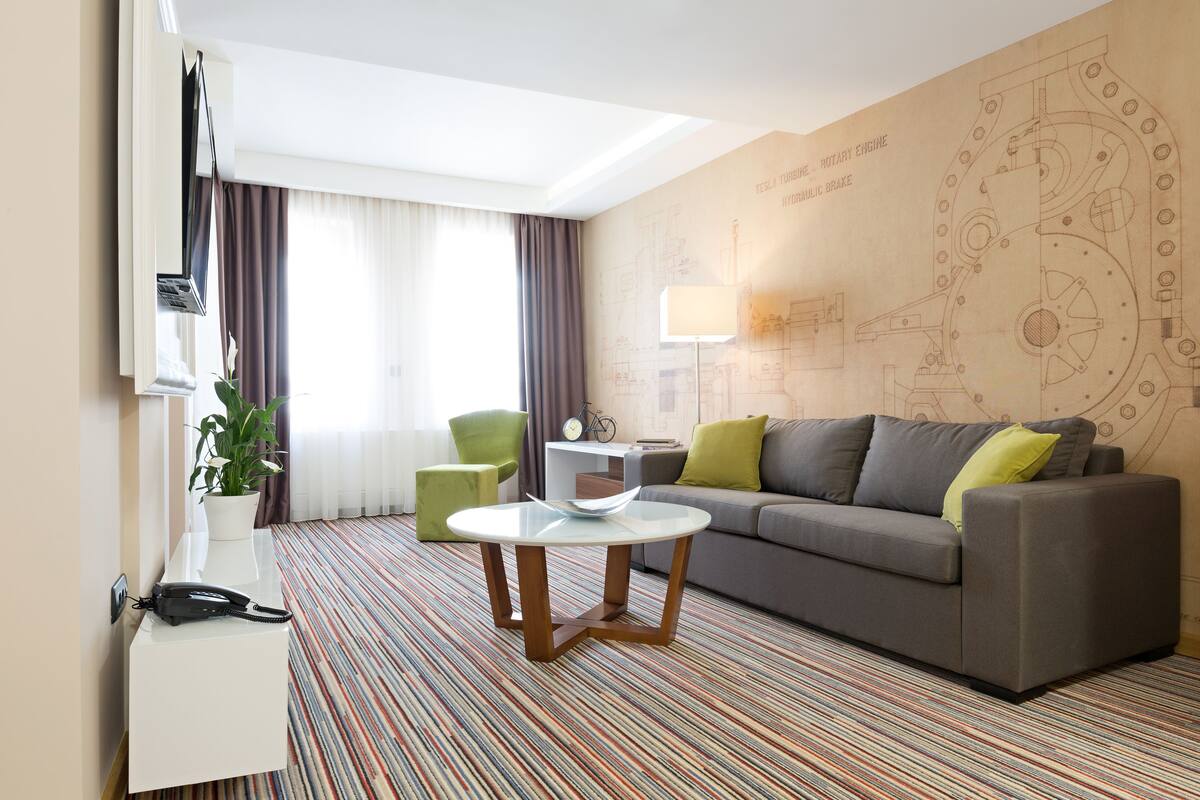 A hotel room with a grey sofa, a small round table, a potted plant, a telephone, and a wall with a mechanical design.