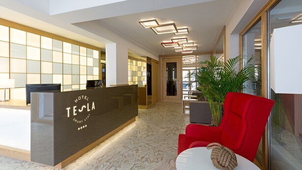 A modern hotel reception area with a Tesla logo, a reception desk, a red chair, and a potted plant. - Hotel Tesla - Smart Stay (Belgrade)