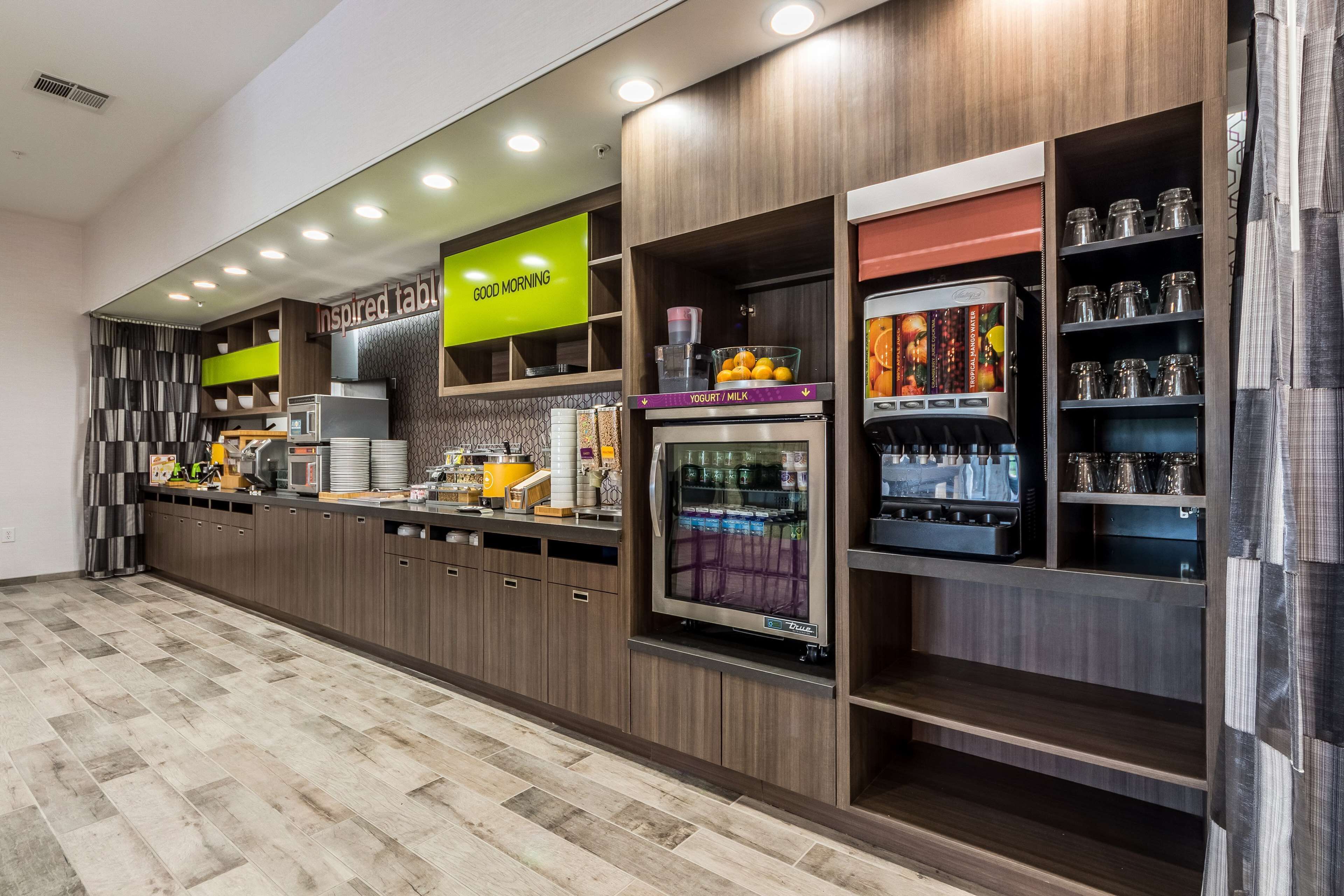 Una barra de desayuno moderna con diversas opciones de comida y bebida, que incluye una estación de yogur y leche, y una máquina de café.
