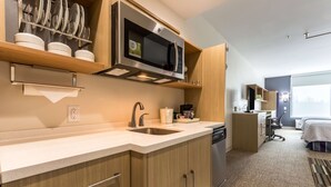 A modern kitchen with a microwave, sink, and wooden cabinets. There is a small dining area with a table and chairs. - Home2 Suites by Hilton Fort Worth Northlake (Roanoke)