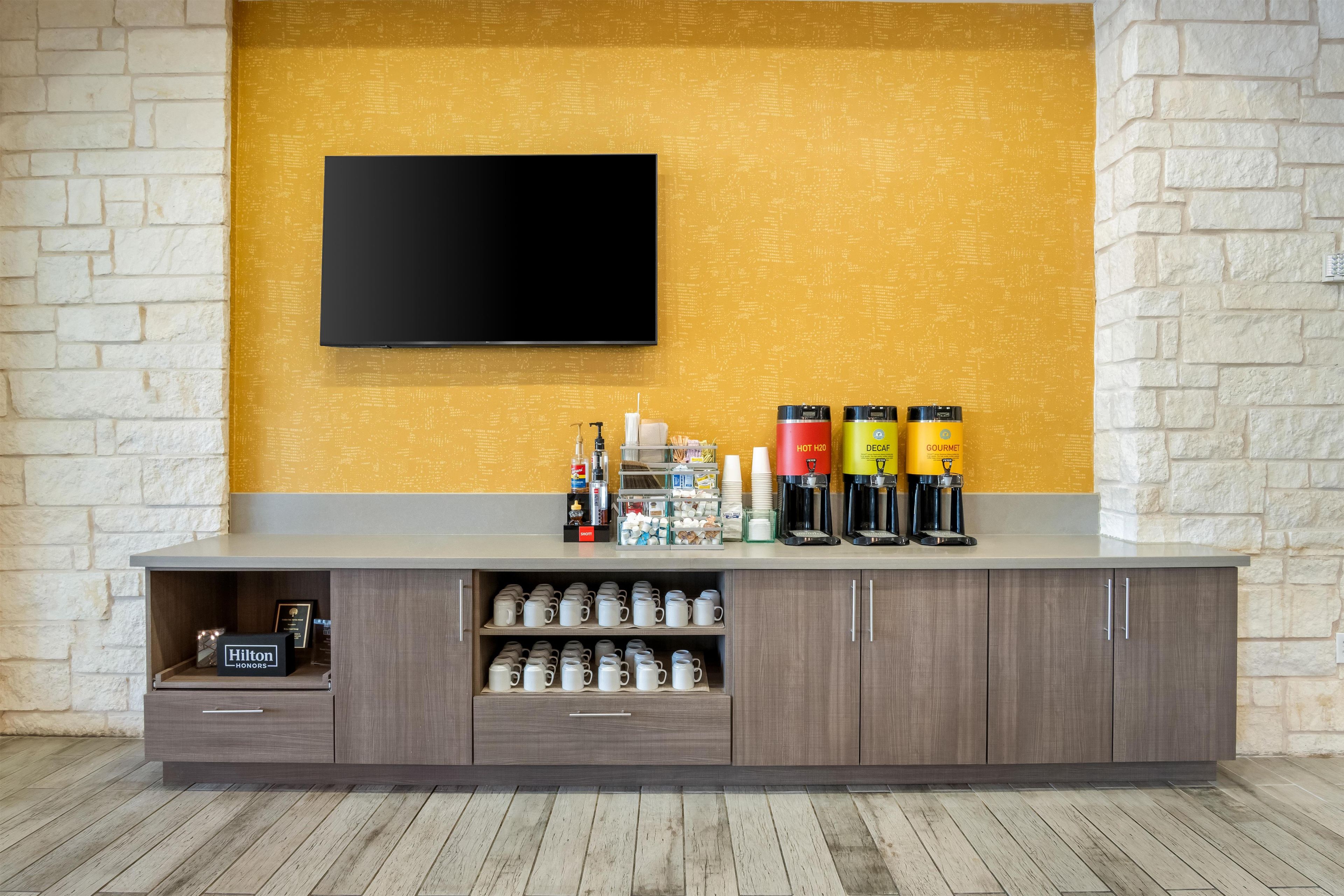 A modern hotel room with a flat-screen TV mounted on a textured yellow wall, a minibar with coffee machines, and a wooden cabinet with storage drawers.