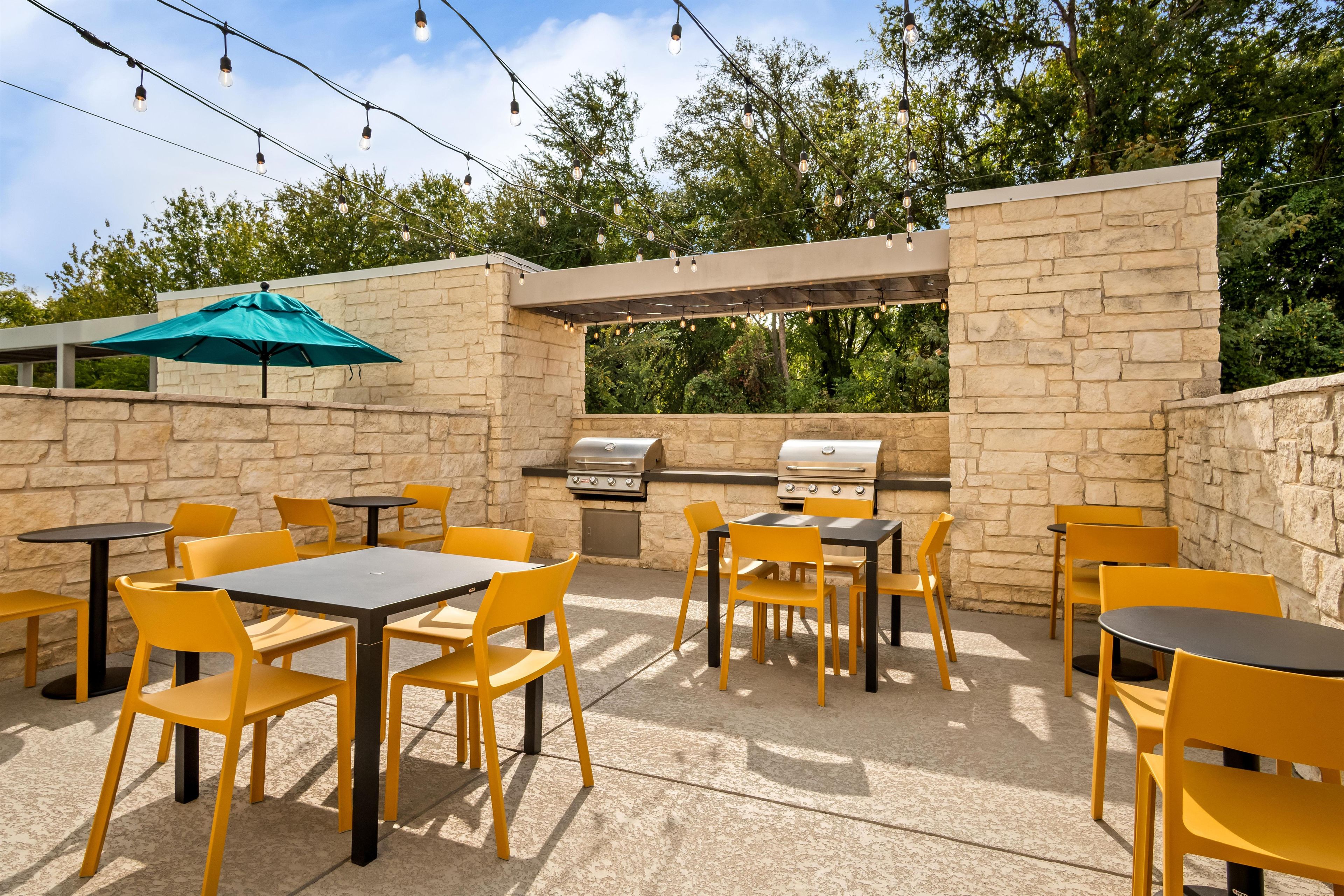 Un patio exterior con muros de piedra, sillas amarillas y mesas, que incluye una parrilla y luces colgantes.