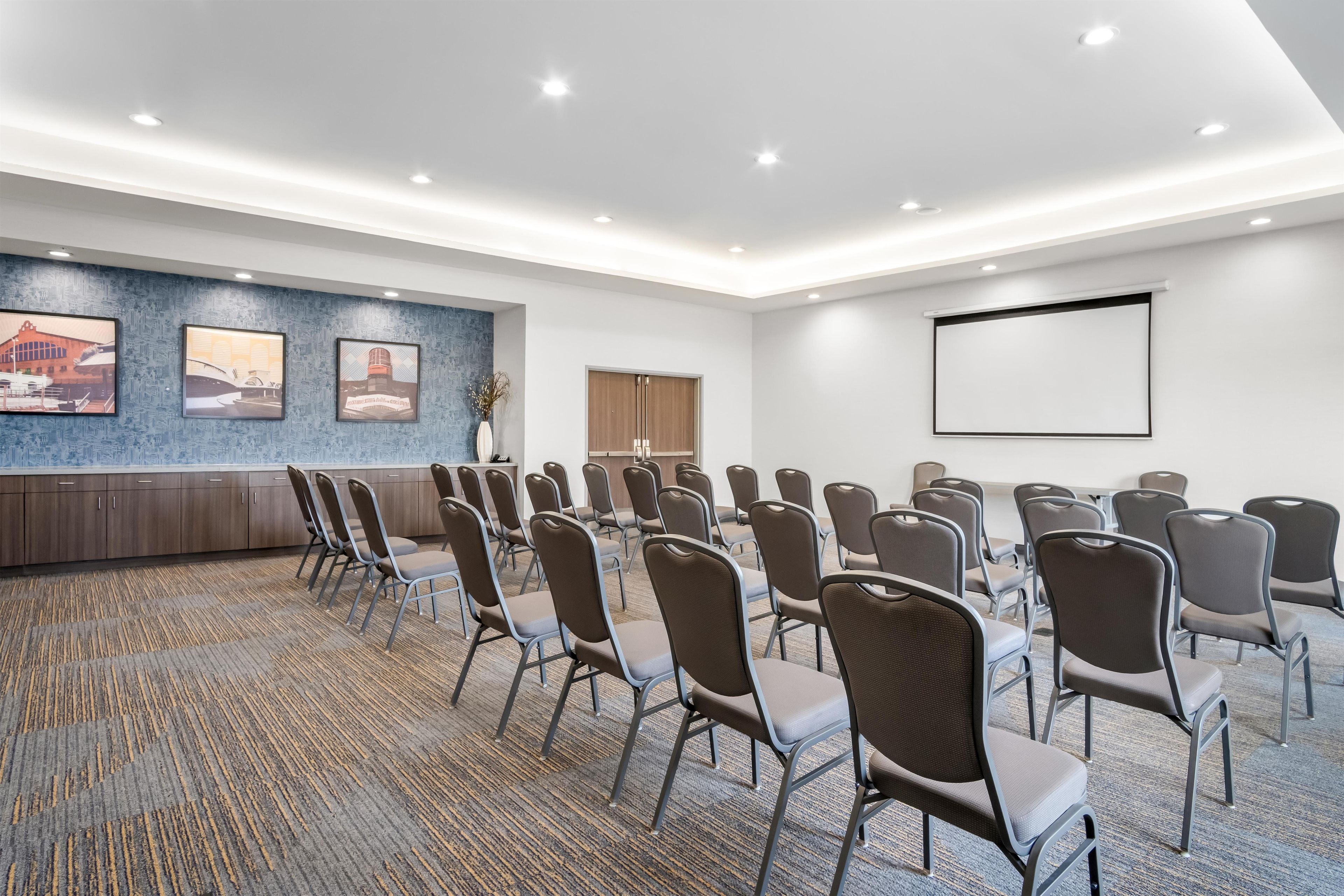 A conference room with rows of chairs, a projector screen, and a carpeted floor.