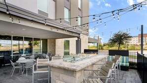 An outdoor patio with stone seating, a glass-top bar, and string lights. - Home2 Suites by Hilton Fort Worth Northlake (Roanoke)