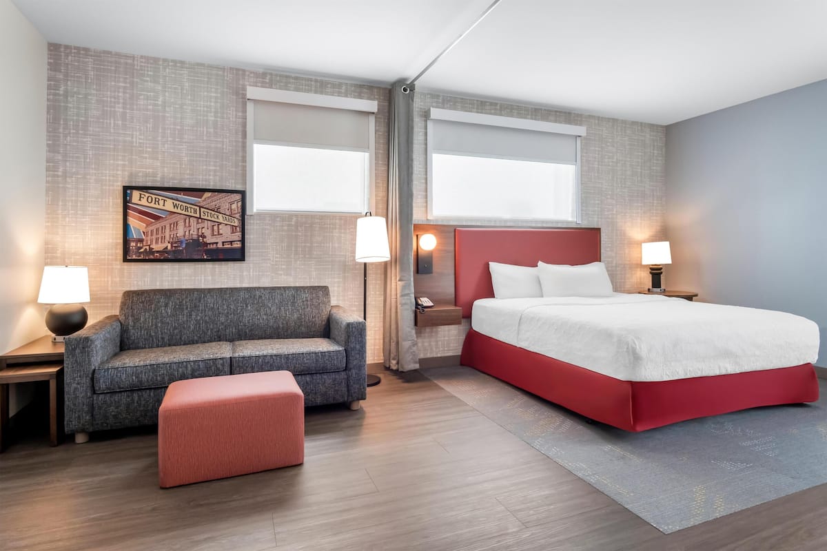Habitación de hotel con cama, sofá, una mesita, lámpara, un cuadro en la pared y ventana.