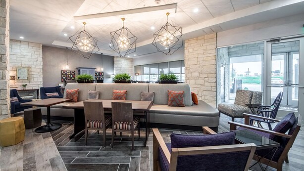 A modern lounge area with a large sofa, dining table, and chairs, featuring geometric pendant lighting and a stone accent wall. - Home2 Suites by Hilton Fort Worth Northlake (Roanoke)