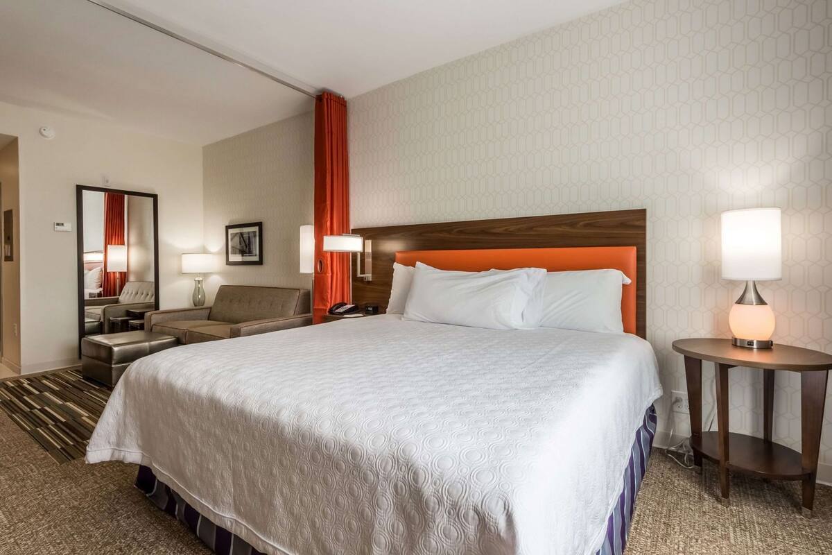 Habitación de hotel con una cama grande, una mesita de noche, una silla, un escritorio y un espejo.