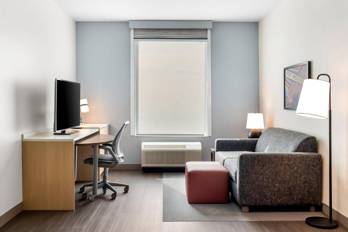 Habitación de hotel con escritorio, silla, televisor, sofá, reposapiés, lámpara de pie y una ventana con persianas.