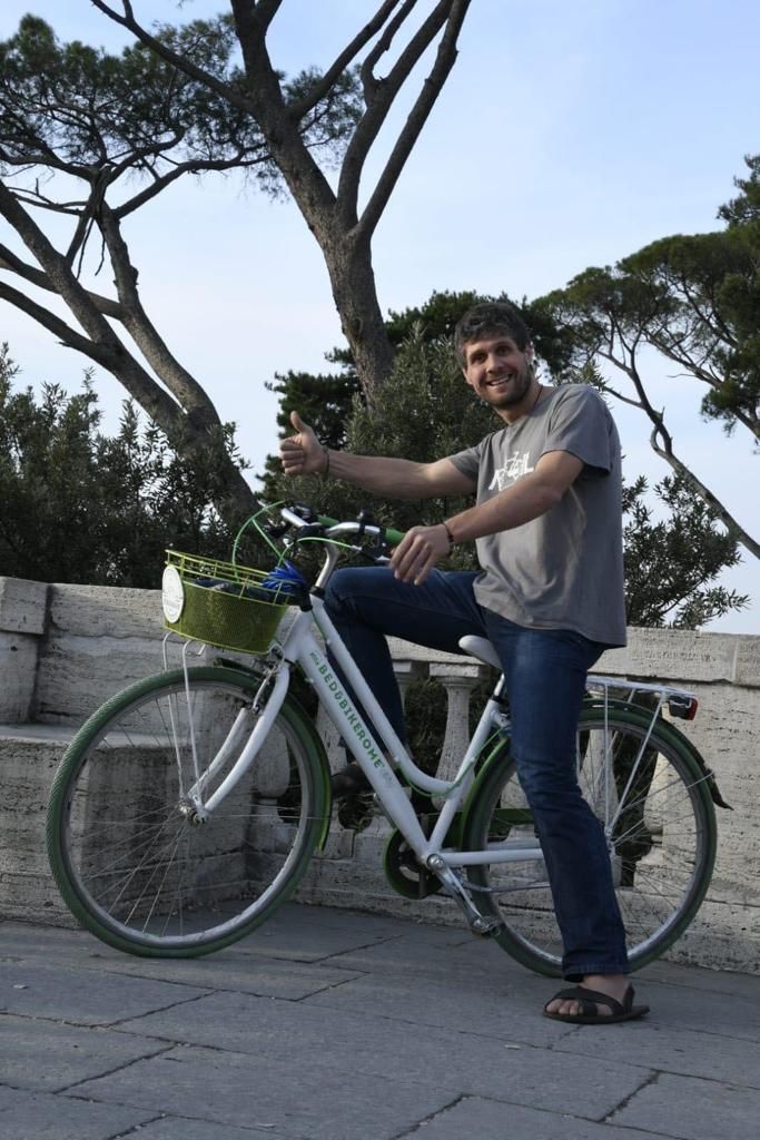 Eine Person sitzt auf einem weißen Fahrrad mit einem grünen Korb, abgestellt auf einer gepflasterten Fläche mit Bäumen im Hintergrund.