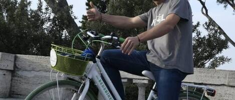 A person sitting on a white bicycle with a green basket, parked on a paved area with trees in the background.