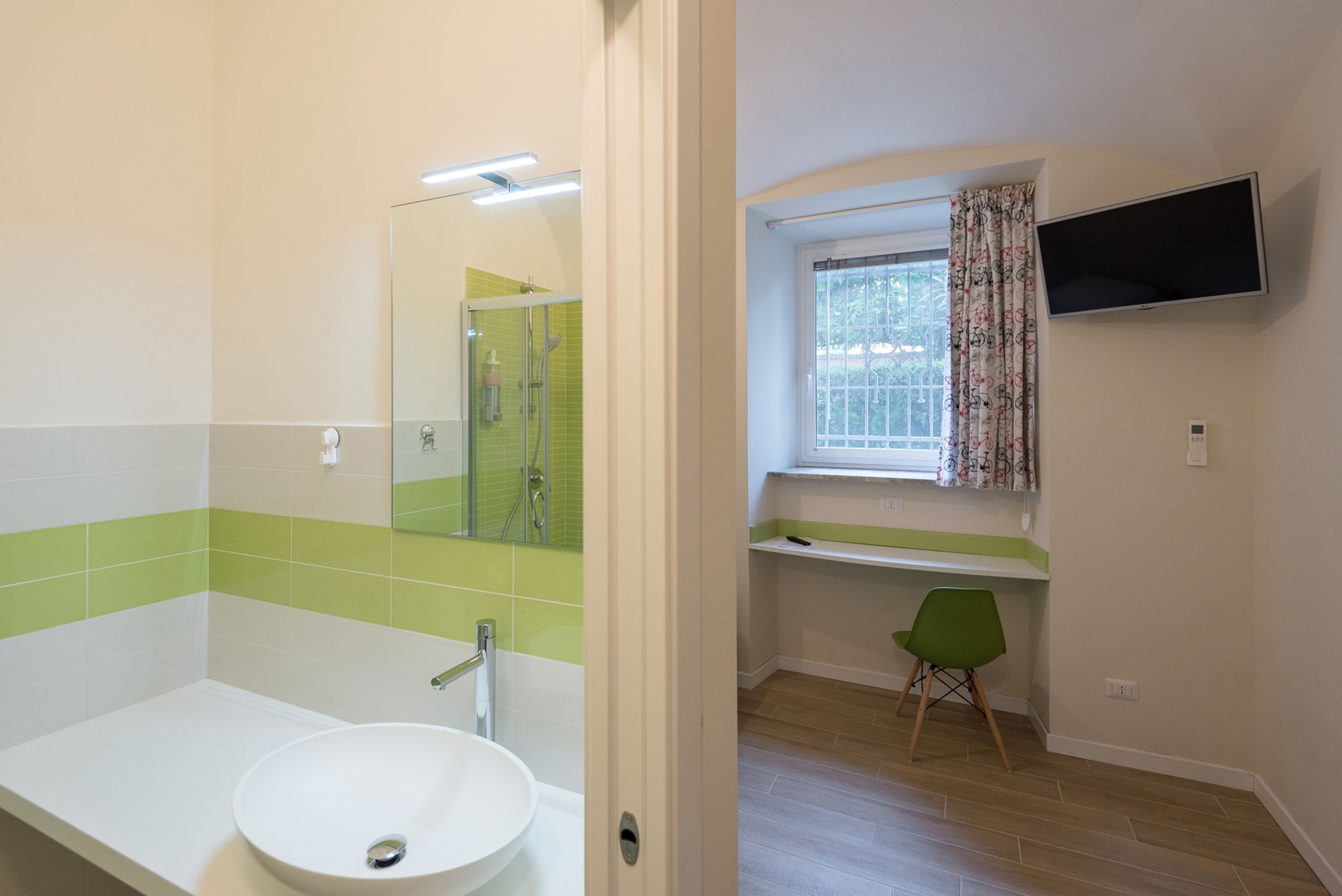 A bathroom with a white sink, a mirror, and a shower. There is a window with curtains and a TV mounted on the wall.
