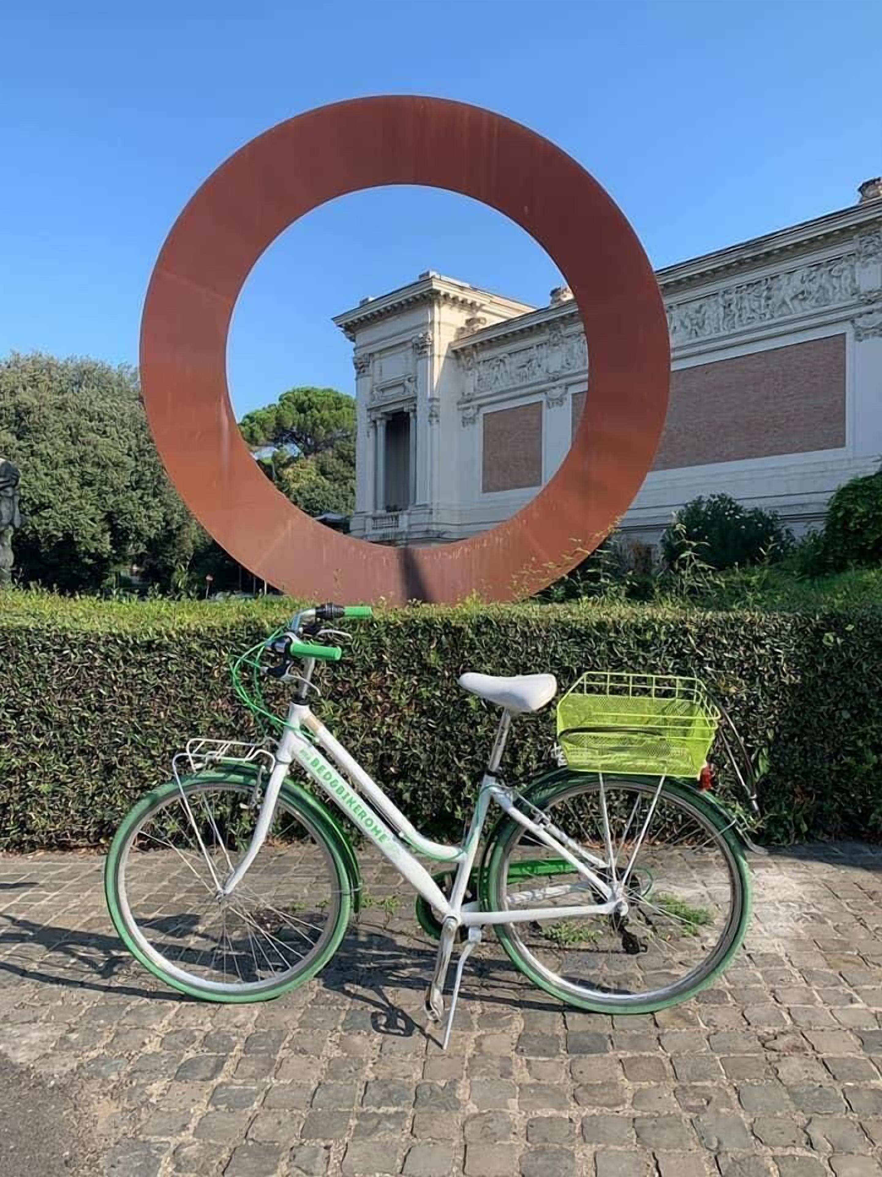 Una bicicleta con una cesta verde estacionada frente a un edificio de arquitectura clásica y una gran escultura circular.