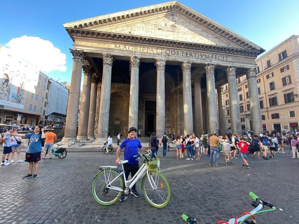 A historic building with classical architecture, including columns and a pediment, in a public square with people and bicycles.