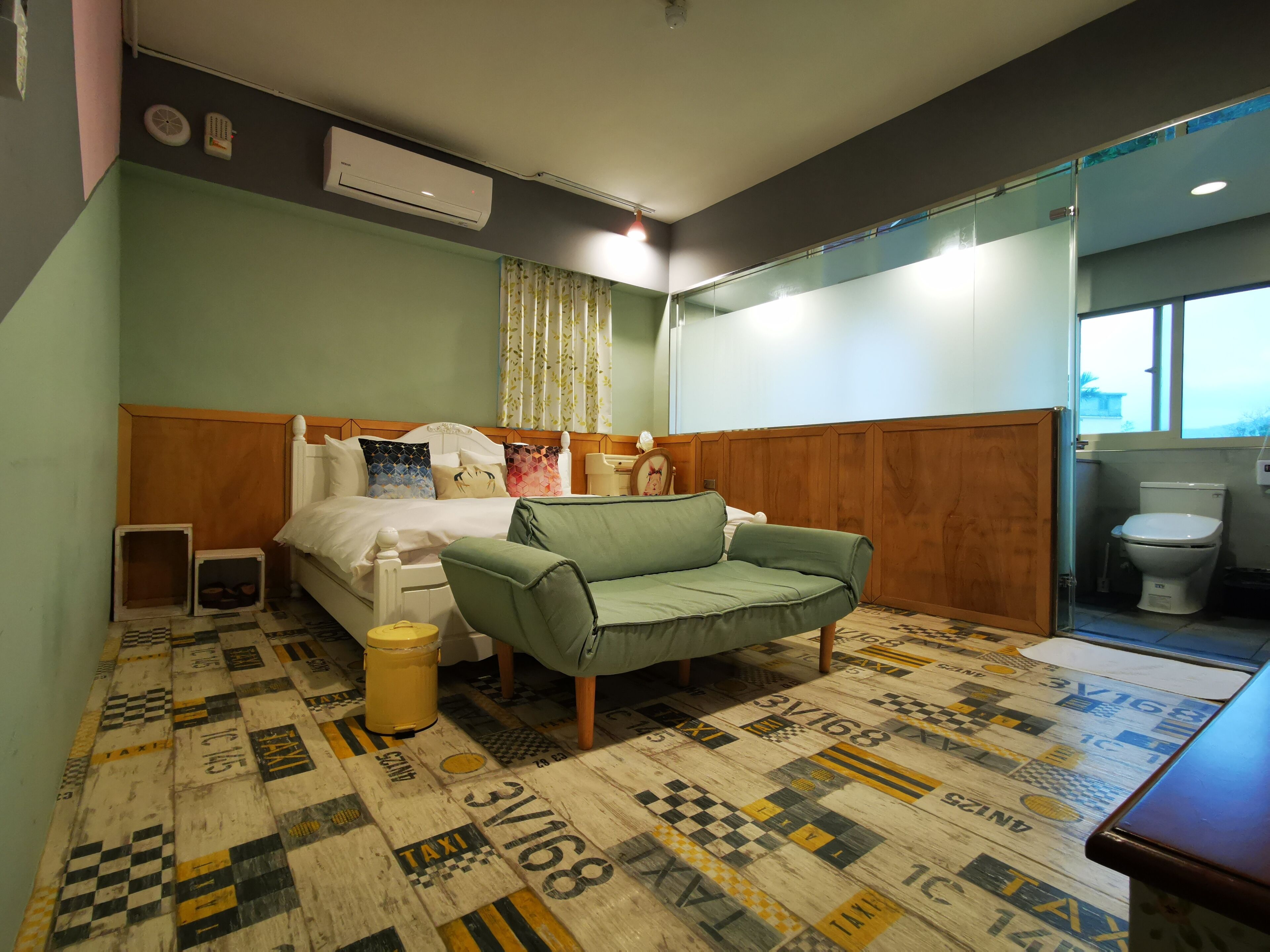 A hotel room with a bed, a green armchair, a small trash bin, and a patterned floor.