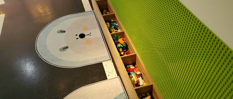 A play area with a green padded wall, a wooden toy storage box, and a floor mat with a bear design.