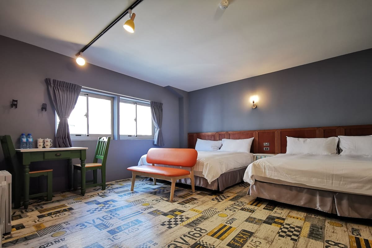 Quarto de hotel com duas camas, uma escrivaninha com cadeiras, uma janela com cortinas e um chão estampado.