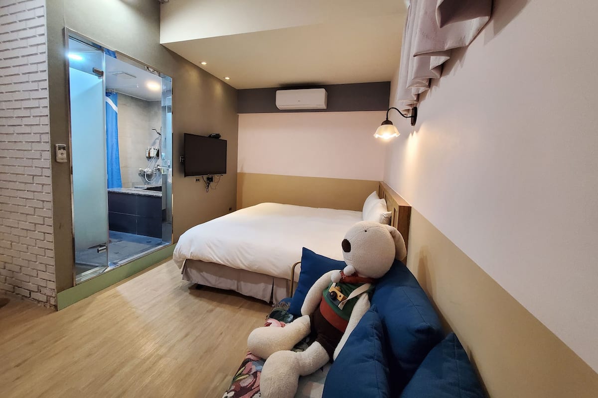 Quarto de hotel com cama, televisão na parede, abajur na mesa de cabeceira e um urso de pelúcia grande sobre a cama.