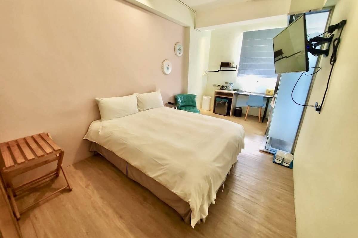 Quarto com cama, um pequeno banco de madeira, escrivaninha com cadeira e televisão na parede.