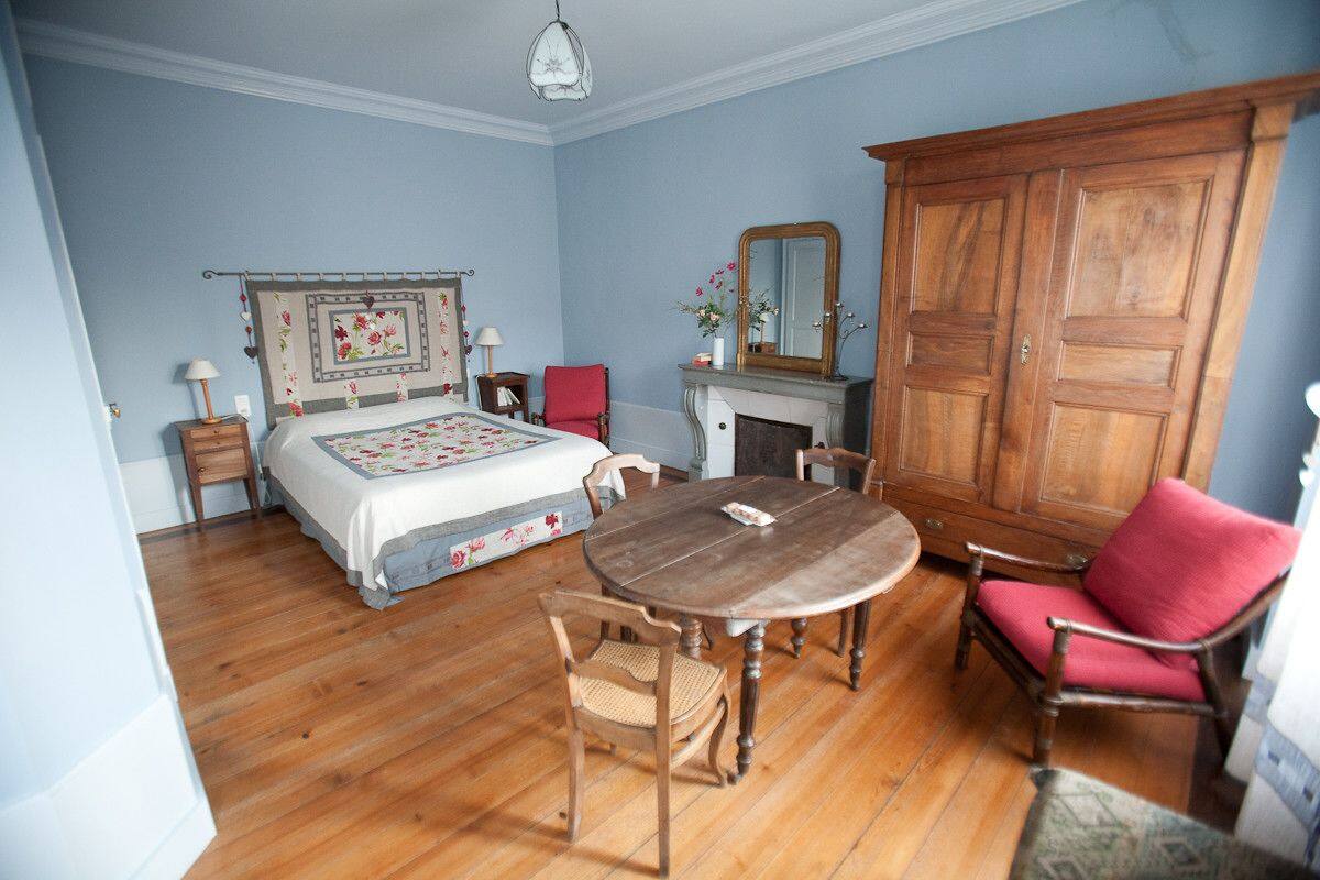 Un dormitorio con cama, una mesa de madera, dos sillas, un sillón rojo, un armario de madera, un espejo y una chimenea.