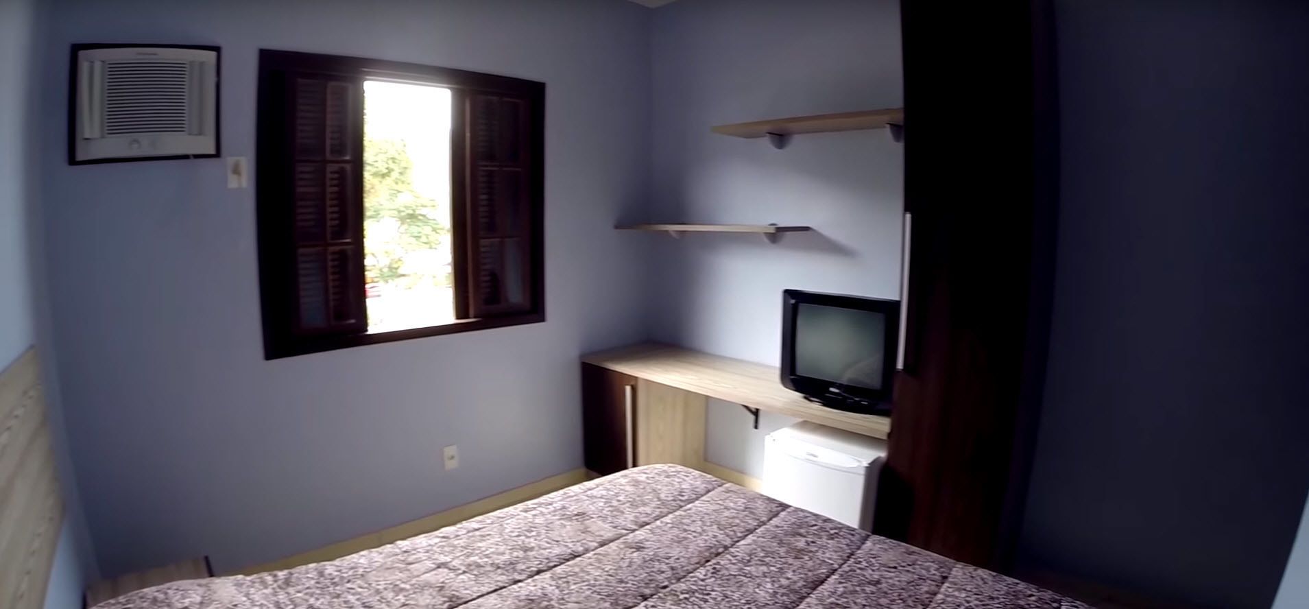 Un dormitorio con una cama, un televisor sobre un mueble, una ventana con contraventanas y una pequeña zona de escritorio.