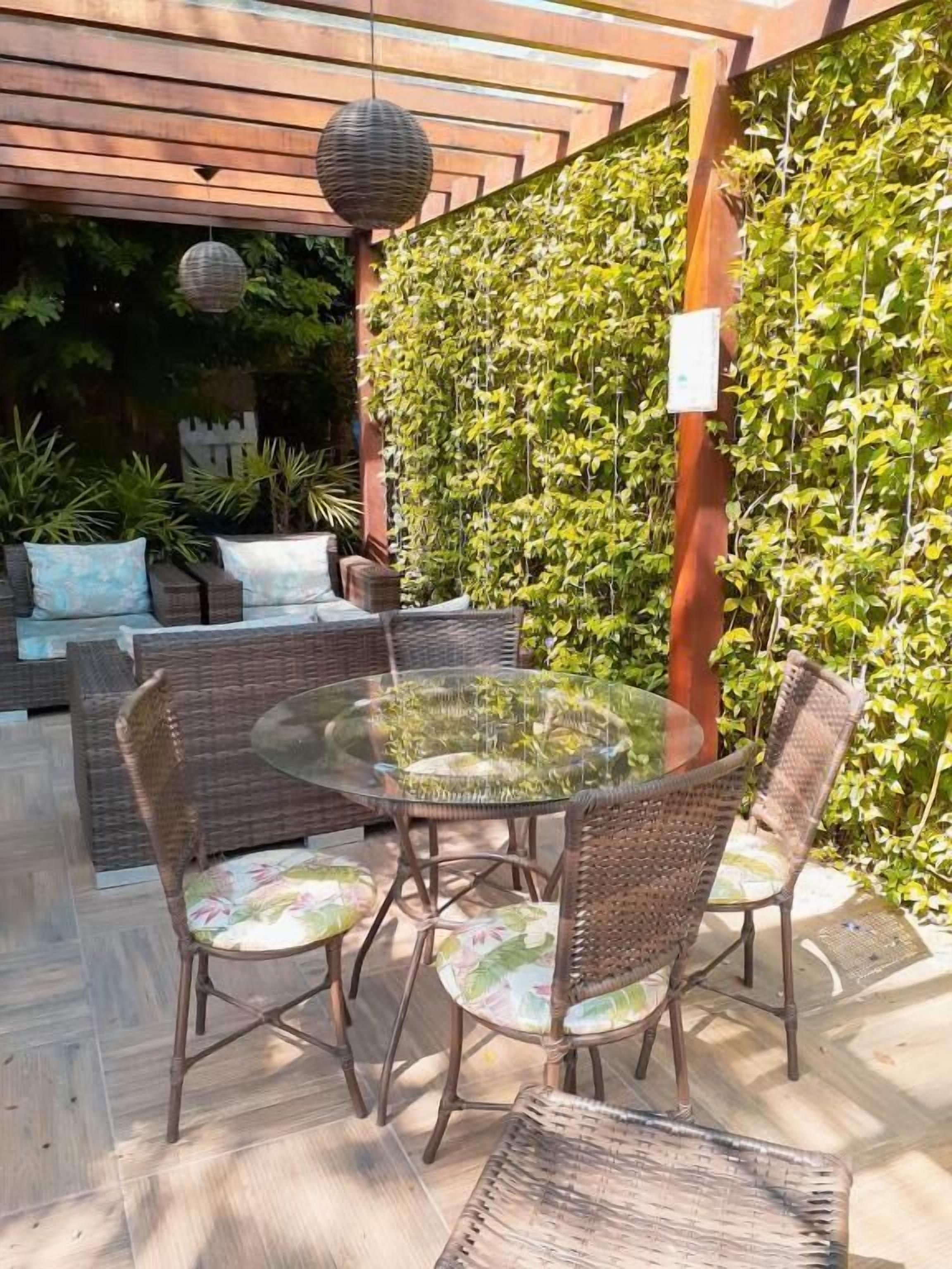 An outdoor patio with wicker furniture and a glass table, surrounded by a wall of climbing plants.