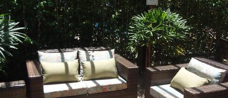 Un sofá de mimbre con cojines, situado en una terraza de madera con un exuberante fondo verde.