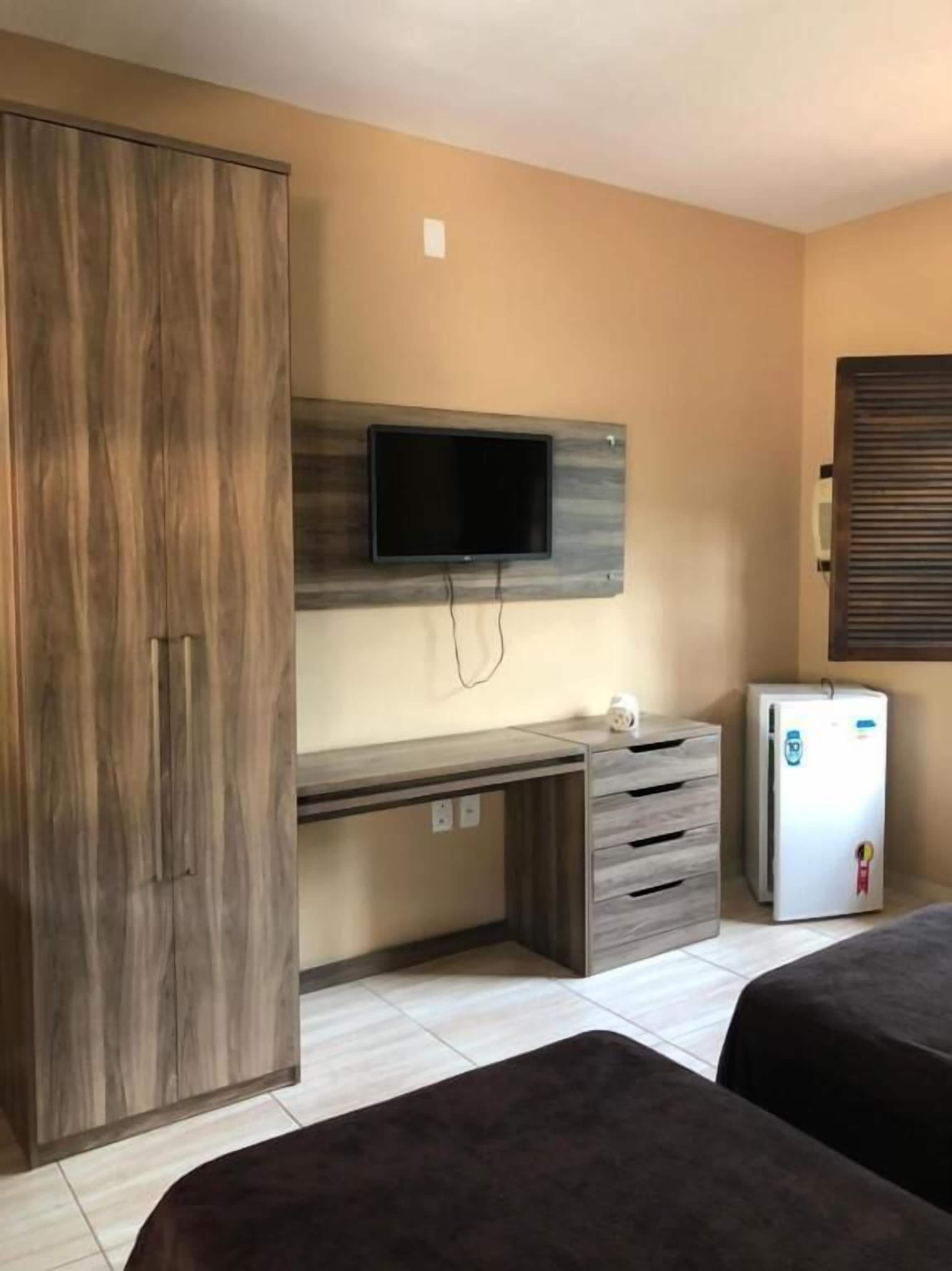 Una habitación con armario, escritorio con cajones, televisor montado en la pared, refrigerador y dos camas.