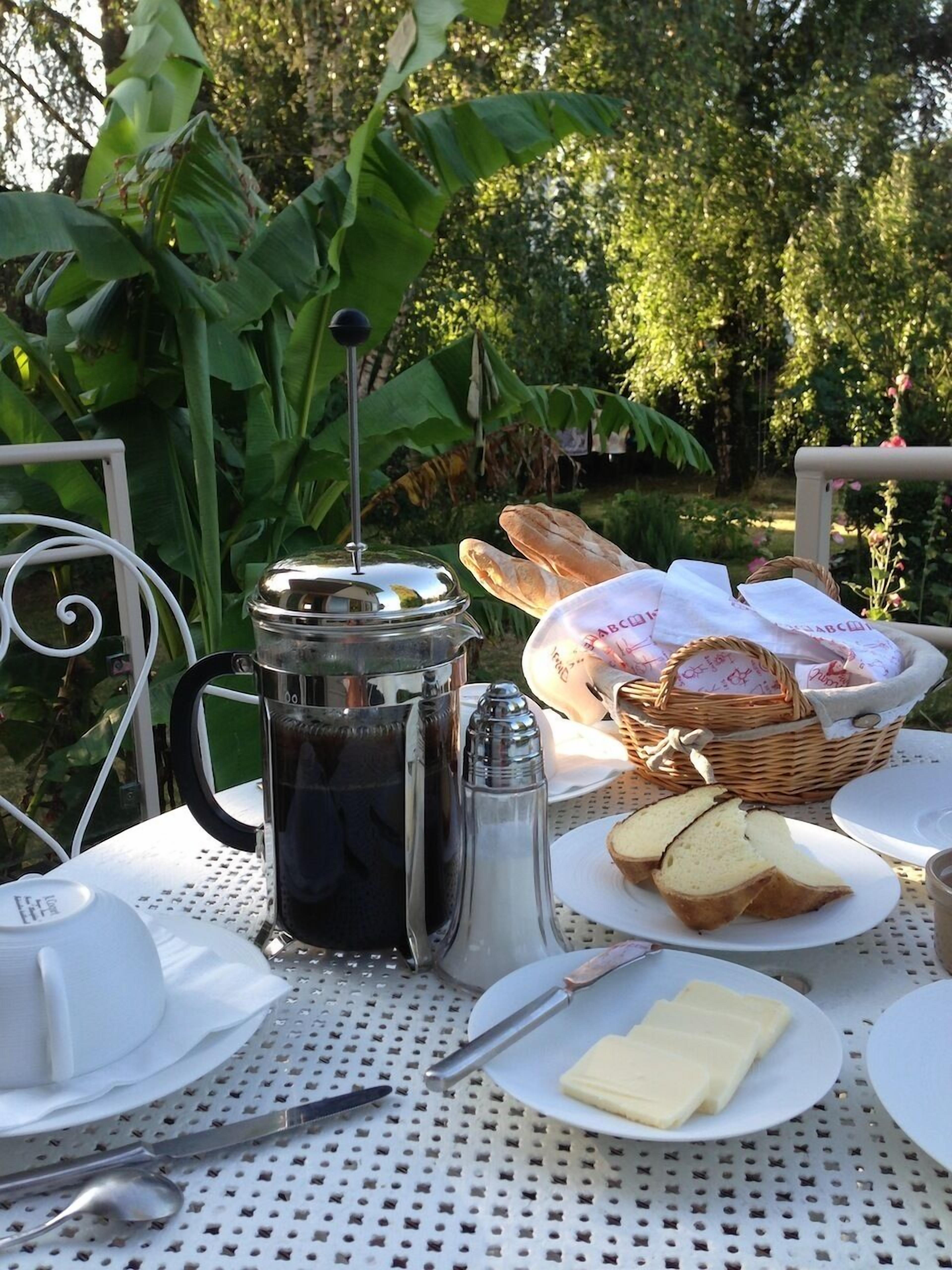 Mesa posta ao ar livre com cafeteira, pão, manteiga e cesta de doces.
