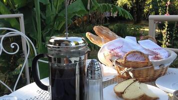Mesa posta ao ar livre com cafeteira, pão, manteiga e cesta de doces.
