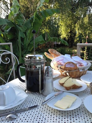 Une table dressée à l’extérieur, avec une cafetière, du pain, du beurre et un panier de viennoiseries.