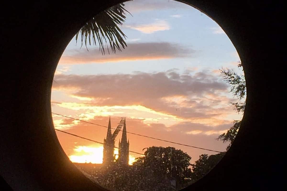 A view through a circular window at sunset, featuring a silhouette of a building with spires and a palm tree.