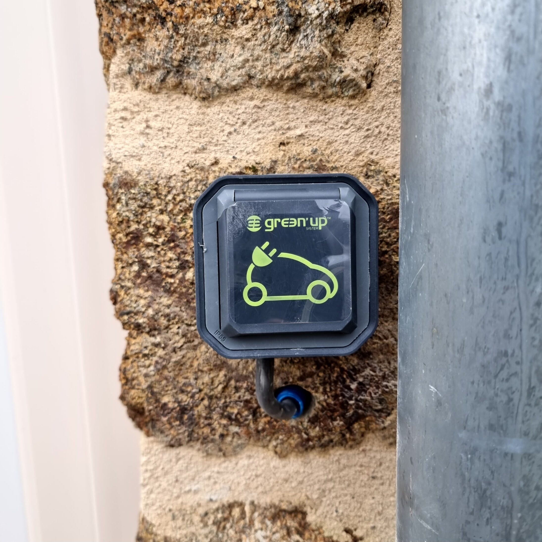 Une borne de recharge pour véhicules  électriques, de couleur noire, avec un logo vert et l’inscription « greenup system » sur un mur texturé.