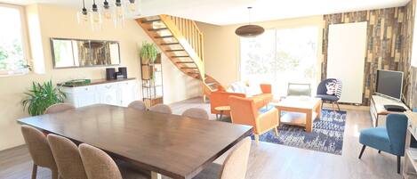 A spacious living area with a wooden dining table, comfortable seating, a staircase, and a large window providing natural light.