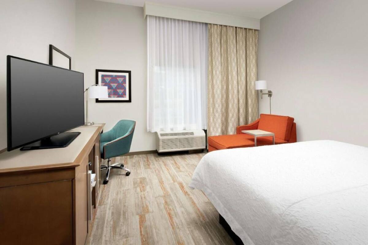 A hotel room with a bed, a desk with a TV, a chair, a small table, and a window with curtains.